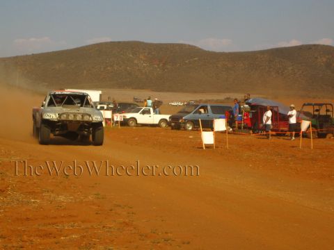 1990 Ford Bronco - number 86 off-road race truck speeds along a dusty red-dirt course at golden hour past spectators, pickups and a course marker.