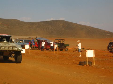 1990 Ford Bronco - number 86 off-road race truck waits on a red-dirt desert course as spectators, a UTV and a flagger stand near course markers.