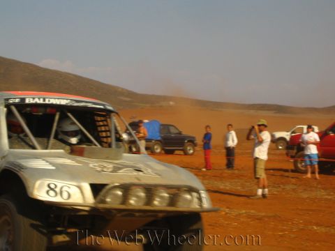 1990 Ford Bronco - white number 86 Baldwin off-road race truck idles on a dusty course as spectators and a photographer watch nearby.