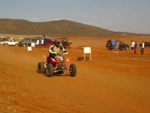 1990 Ford Bronco - helmeted rider races a red ATV quad across a red-dirt desert course past spectators, pickups and course markers.
