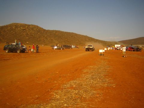 1990 Ford Bronco - off-road race truck kicks up dust on a wide red-dirt desert course lined with spectators and parked vehicles at golden hour.