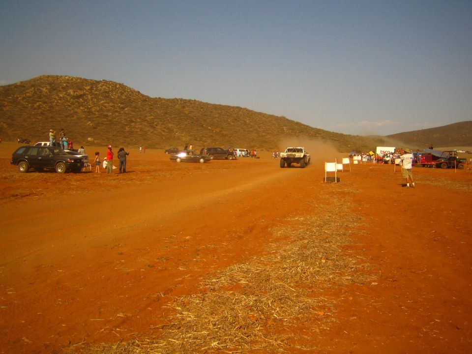 Off-road race truck on a wide red dirt course with spectators at golden hour