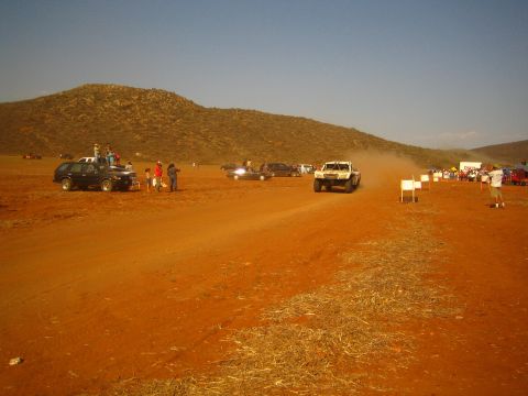 1990 Ford Bronco - light-colored off-road race truck speeds along a red-dirt desert course at sunset past parked vehicles and spectators.
