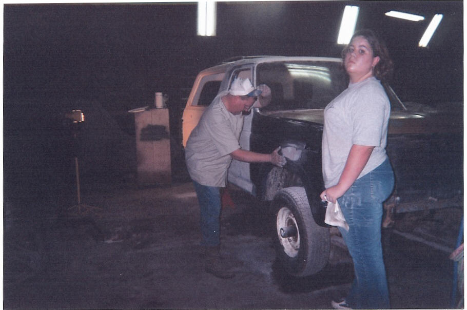 Two people working on a light-colored truck in a dark garage, 3/4 rear-side view