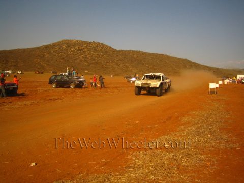 1990 Ford Bronco - off-road race truck races across a red-dirt desert course raising dust as spectators and parked trucks line the marked route.
