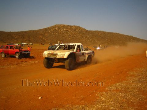 1990 Ford Bronco - white and blue off-road race truck speeds along a red-dirt desert course past a red SUV and spectators raising a trail of dust.