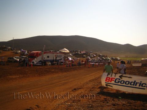 1990 Ford Bronco - BF Goodrich race pit banner marks a busy desert pit area with hauler trucks, tents and a crowd gathered below a hill at dusk.