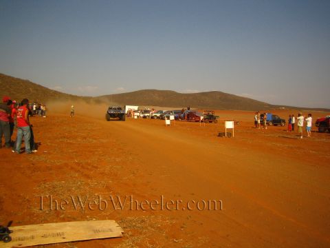 1990 Ford Bronco - Spectators stand beside parked vehicles along a red-dirt desert track while a race truck raises dust near distant hills.