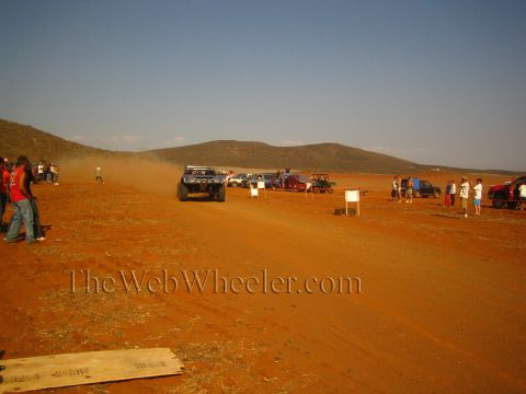 1990 Ford Bronco - race truck barrels down a red-dirt desert course trailing dust as spectators and parked trucks watch from the side.