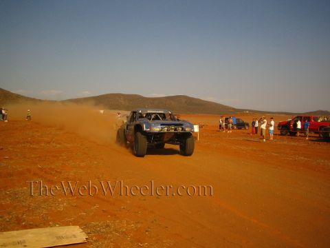 1990 Ford Bronco - open-wheel off-road race truck launches over a red-dirt course in a dust cloud as spectators look on.