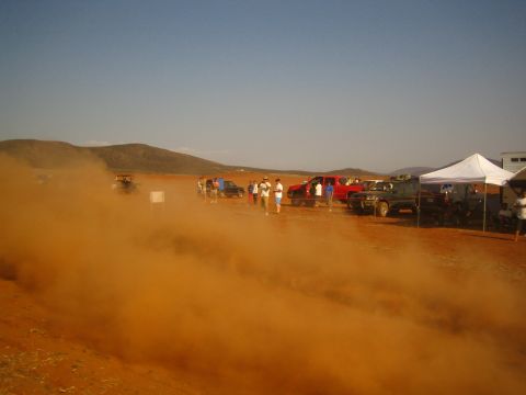 1990 Ford Bronco - thick orange dust cloud drifts across a desert pit area dotted with parked trucks, spectators and a white pop-up tent.