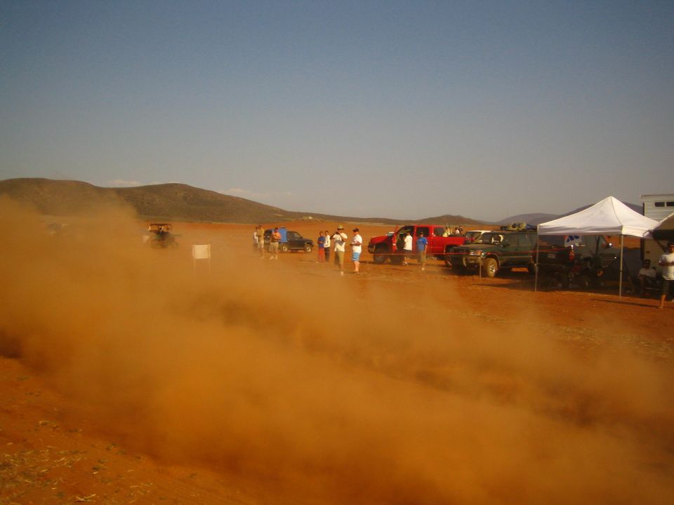 Thick orange dust cloud over a desert pit area with parked trucks and a white tent
