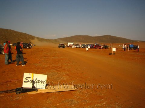 1990 Ford Bronco - fallen white 'Safari' sign lies in red dirt in the foreground of a desert race course lined with parked vehicles and spectators.