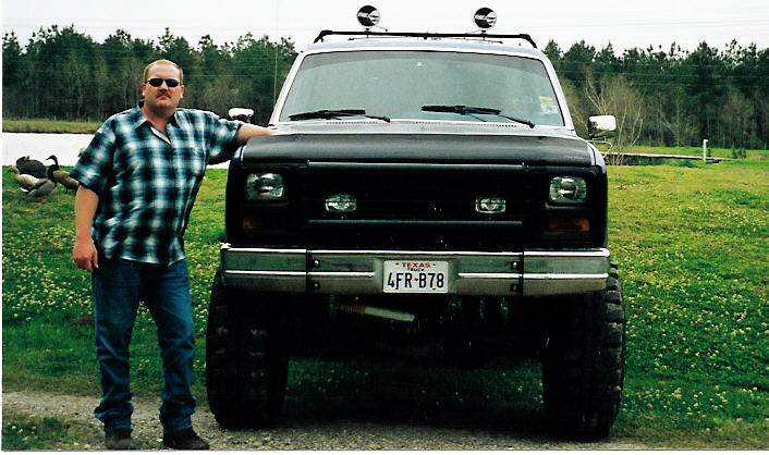 Man in plaid shirt standing beside a lifted black-and-white Ford Bronco with roof lights and Texas plate, front view