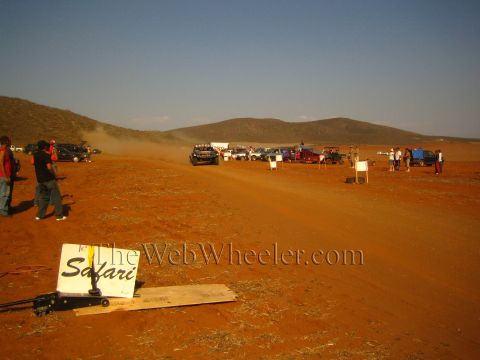 1990 Ford Bronco - 'Safari' sign rests in the red dirt as race vehicles and spectators gather along a dusty desert course toward distant hills.