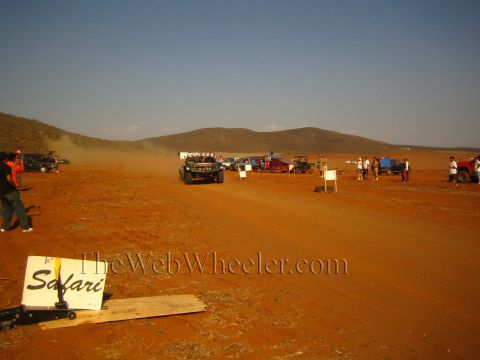 1990 Ford Bronco - off-road buggy travels a red-dirt desert course trailing dust, with a fallen 'Safari' sign in the foreground and spectators lining the rout…