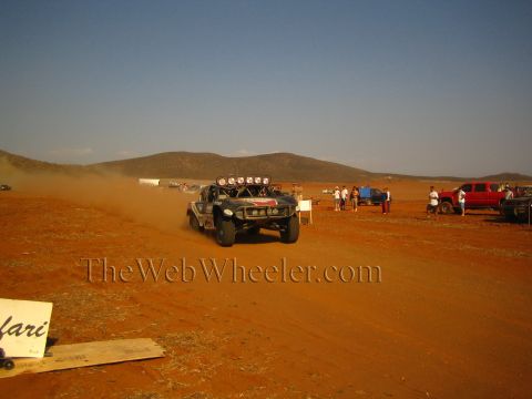 1990 Ford Bronco - black off-road race truck fitted with roof lights speeds down a red-dirt course trailing dust as spectators and trucks watch from the side.