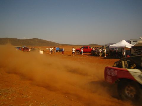 1990 Ford Bronco - red off-road truck throws up heavy dust passing a desert pit area with a white tent, parked pickups and spectators.