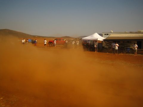 1990 Ford Bronco - Orange dust billows across a desert pit area where spectators gather near a white tent, a building and parked trucks.