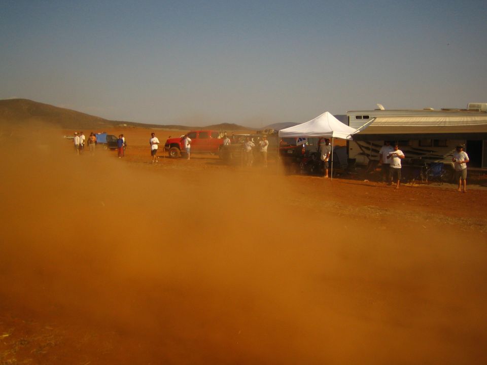 Orange dust billows across a desert pit with a white tent, building and parked trucks