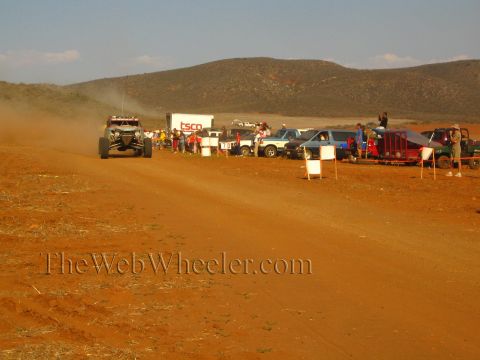 1990 Ford Bronco - off-road buggy runs a red-dirt desert course past a 'TSCO' box trailer, parked vehicles and a crowd of spectators.