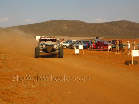 1990 Ford Bronco - open-wheel off-road race truck travels a red-dirt course beside parked pickups, a UTV and spectators in the desert.