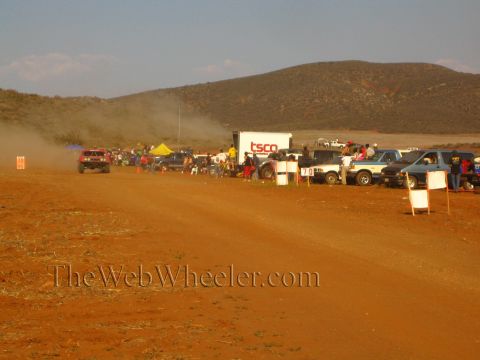 1990 Ford Bronco - Spectators and parked trucks line a red-dirt desert course near a white 'TSCO' box trailer as a vehicle raises dust.