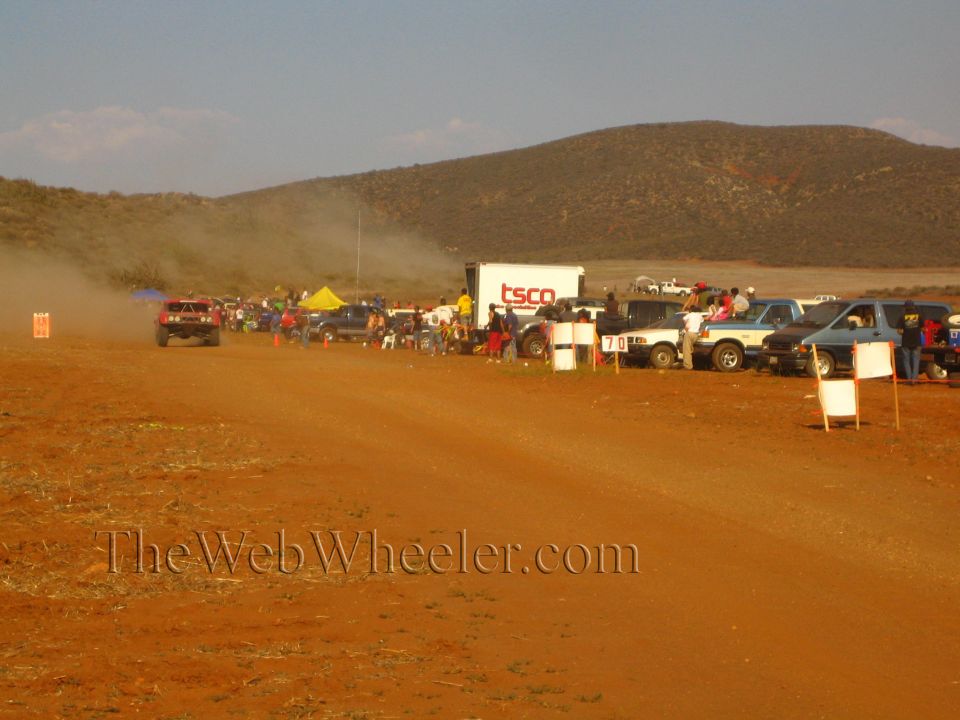 Crowd and parked trucks line a desert course near a white 'TSCO' trailer