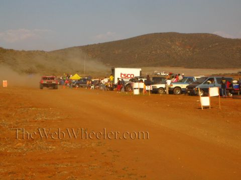 1990 Ford Bronco - row of parked trucks and spectators lines a red-dirt desert course beside a white 'TSCO' trailer under distant hills.