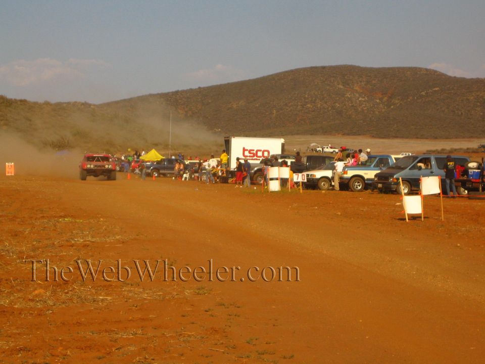 Pit and spectator area along a desert course with a 'TSCO' trailer and parked trucks