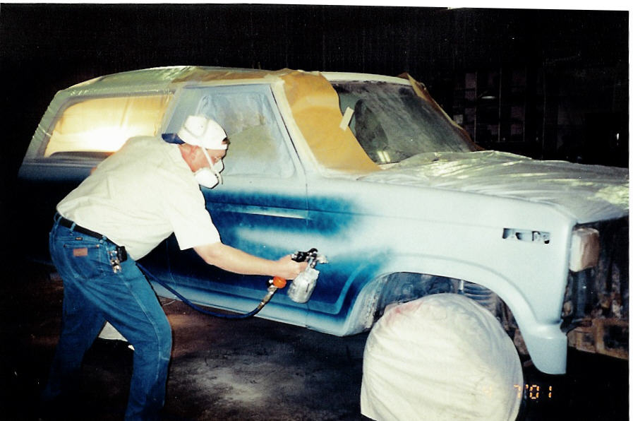 Man in respirator spray-painting side of light blue Ford Bronco in a shop