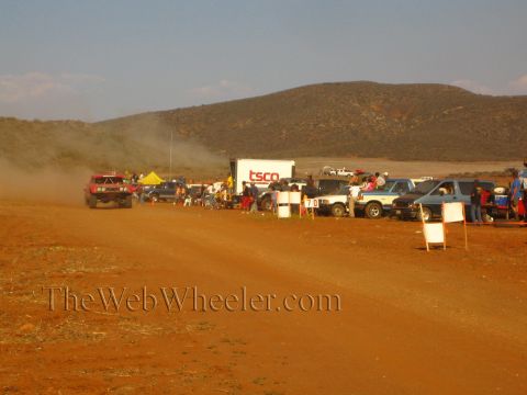 1990 Ford Bronco - red off-road truck runs a red-dirt course past a white 'TSCO' trailer, parked trucks and a roadside crowd of spectators.
