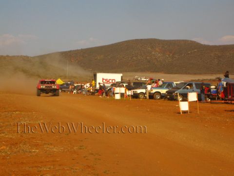1990 Ford Bronco - red off-road truck speeds down a red-dirt desert course past a 'TSCO' trailer, a line of parked vehicles and spectators.