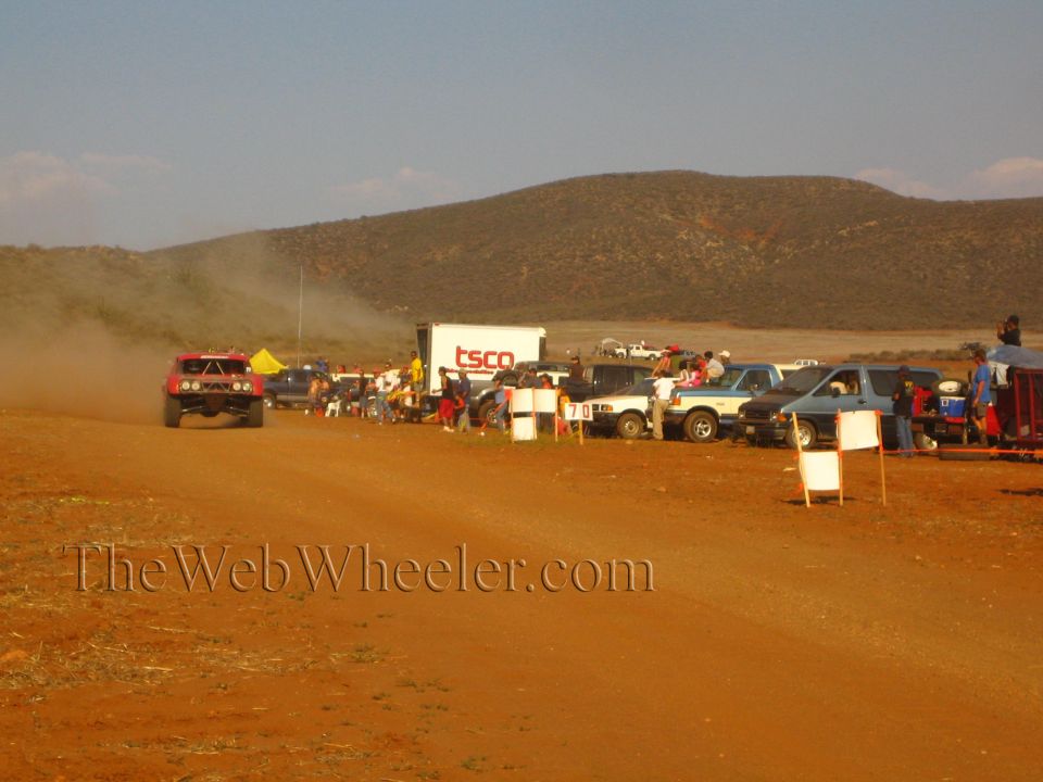 Red off-road truck speeds a dirt course past a 'TSCO' trailer and parked vehicles