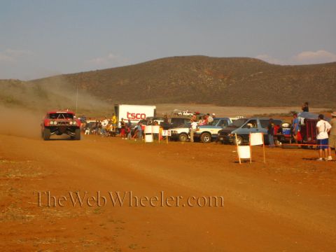 1990 Ford Bronco - red off-road truck travels a red-dirt course past a 'TSCO' trailer as spectators watch from behind a roped-off pit line.
