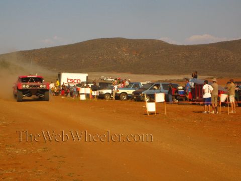 1990 Ford Bronco - red off-road truck races a red-dirt course beside a 'TSCO' trailer, parked pickups and spectators under desert hills.