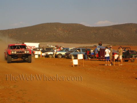 1990 Ford Bronco - red off-road truck runs a red-dirt course past a 'TSCO' trailer, parked trucks, a UTV and spectators behind a rope line.