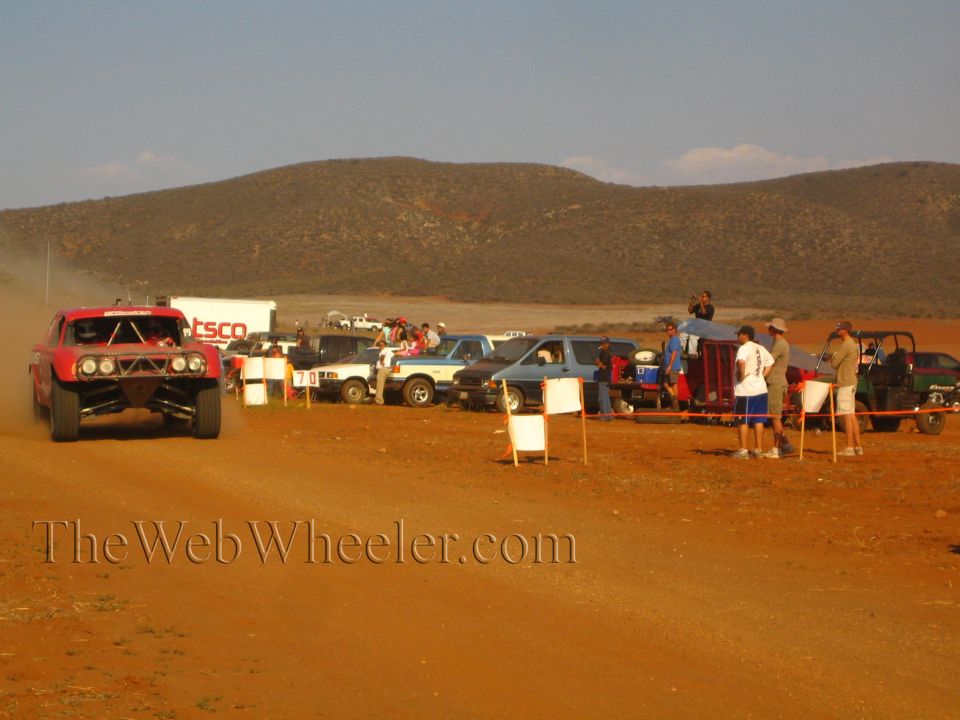 Red off-road truck on a dirt course past a 'TSCO' trailer, parked trucks and a UTV