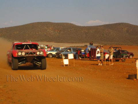 1990 Ford Bronco - red off-road truck speeds along a red-dirt desert course past parked trucks, a green UTV and spectators behind a rope.