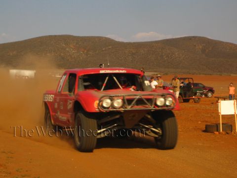 1990 Ford Bronco - red off-road truck with round headlights and a tube front end powers down a red-dirt desert course trailing dust past spectators.