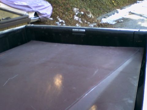 1973 Ford Ranchero - The bed of a pickup truck is covered by a tonneau cover, with a portion of another vehicle covered by a tarp in the background.
