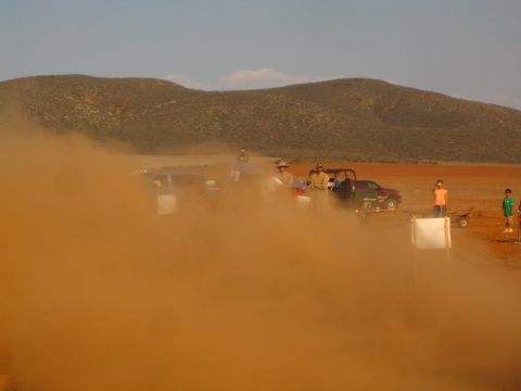 1990 Ford Bronco - thick orange dust cloud drifts over a desert pit area where parked trucks, spectators and a child in orange gather.