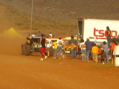 1990 Ford Bronco - crowd of people swarms race vehicles in a dusty desert pit beside a white 'TSCO' box trailer.