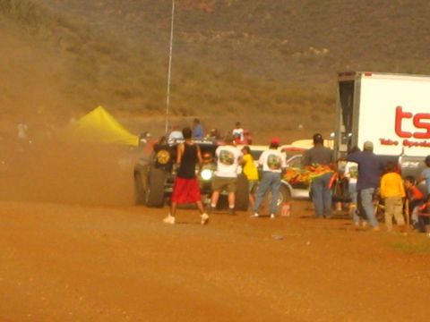 1990 Ford Bronco - People crowd around off-road race trucks in a dusty red-dirt pit beside a white 'TSCO' trailer and a yellow tent.