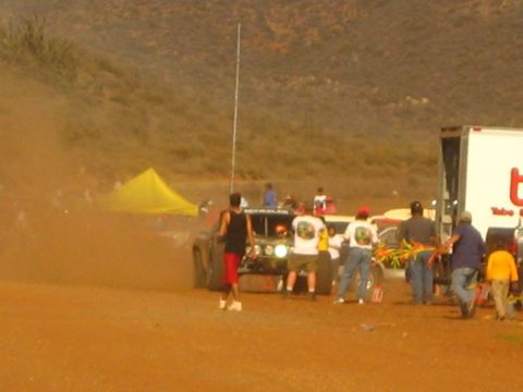 1990 Ford Bronco - Spectators stand near an off-road race truck kicking up red dust beside a box trailer in the desert.