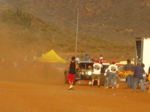 1990 Ford Bronco - dark off-road race truck idles amid spectators and a yellow canopy in a dusty desert pit.