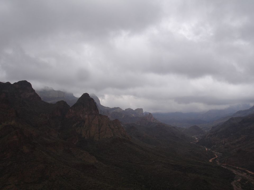 Mountain valley with a winding dirt road under a cloudy sky. Supermotors.net logo in lower right
