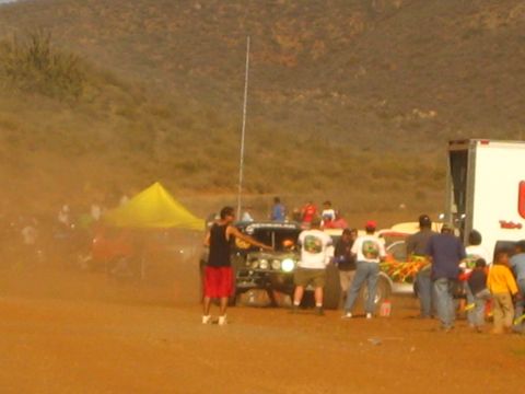 1990 Ford Bronco - crowd gathers around an off-road race truck stirring red dust near a white box trailer.