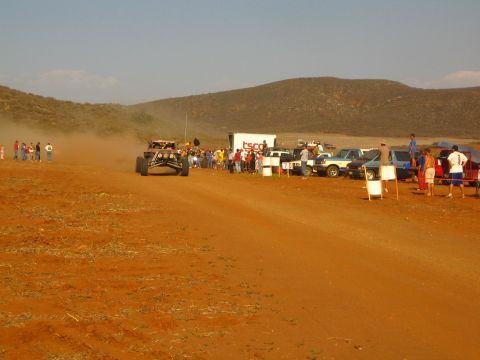 1990 Ford Bronco - off-road race buggy speeds along a red dirt course as spectators line up by parked vehicles.