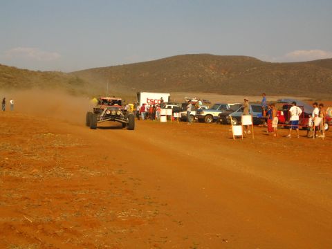 1990 Ford Bronco - red off-road buggy trails dust toward spectators and parked trucks on a desert race course.